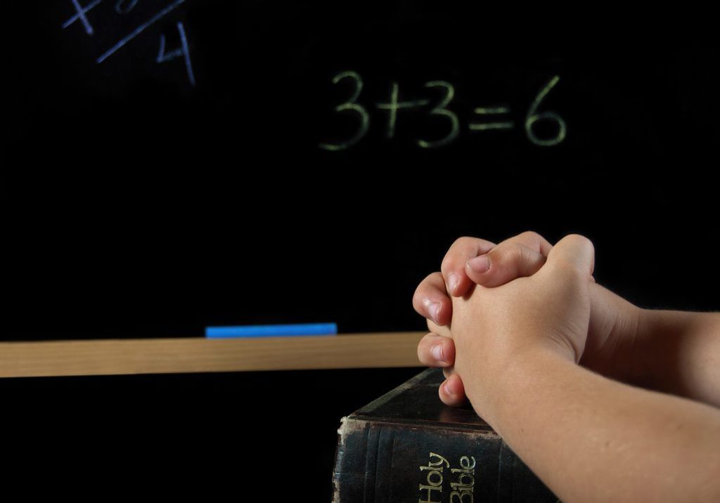 Child's hand folded in prayer on a Holy Bible in a classroom with chalkboard.